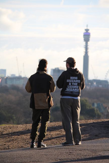 Two individuals are standing outdoors on a rooftop terrace, with City Hall and the BT Tower visible in the background under a partly cloudy sky. The person on the left has dark hair and is wearing a t