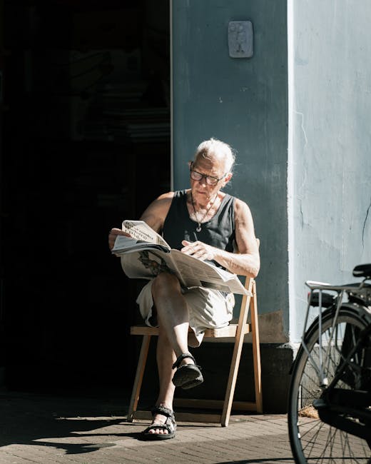 An elderly woman with short gray hair and glasses is seated outdoors on a wooden chair, reading a newspaper. She is wearing a sleeveless black top, beige shorts, and black sandals. The scene is set ag